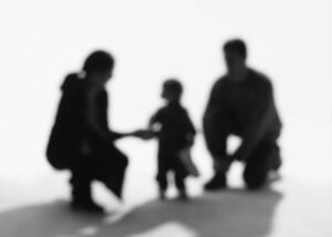 The image shows a blurred silhouette of three people. Two adults, one on the left and one on the right, are crouching or kneeling and facing each other. A small child stands between them, holding hand of one adult. The background is bright, making the figures appear dark and out of focus.